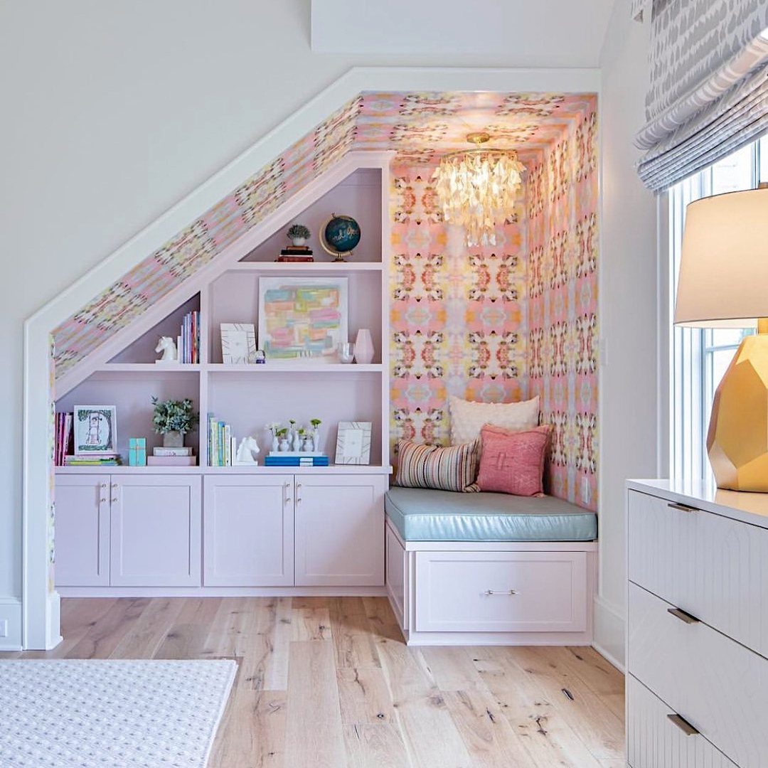 Built-in reading nook under a staircase with lavender cabinets, patterned wallpaper, and soft cushions.