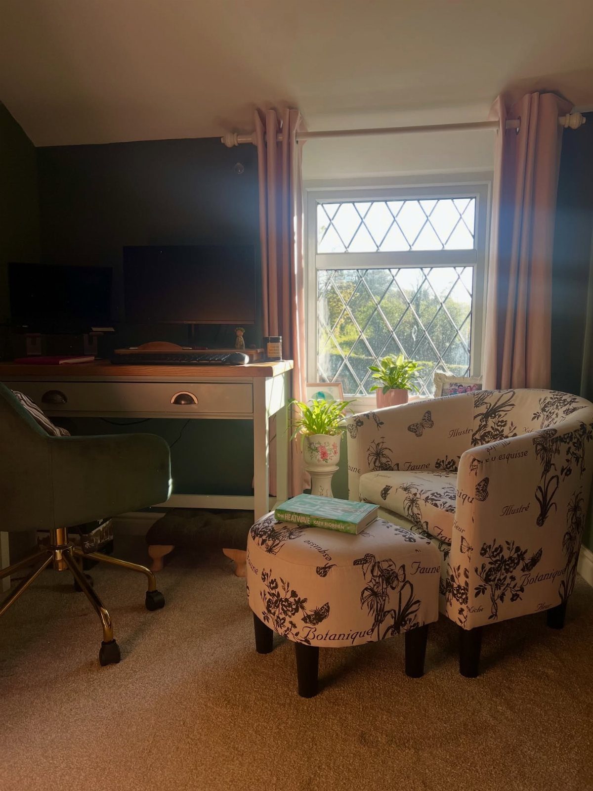 Patterned armchair and ottoman positioned near a window with pink curtains and small plants.