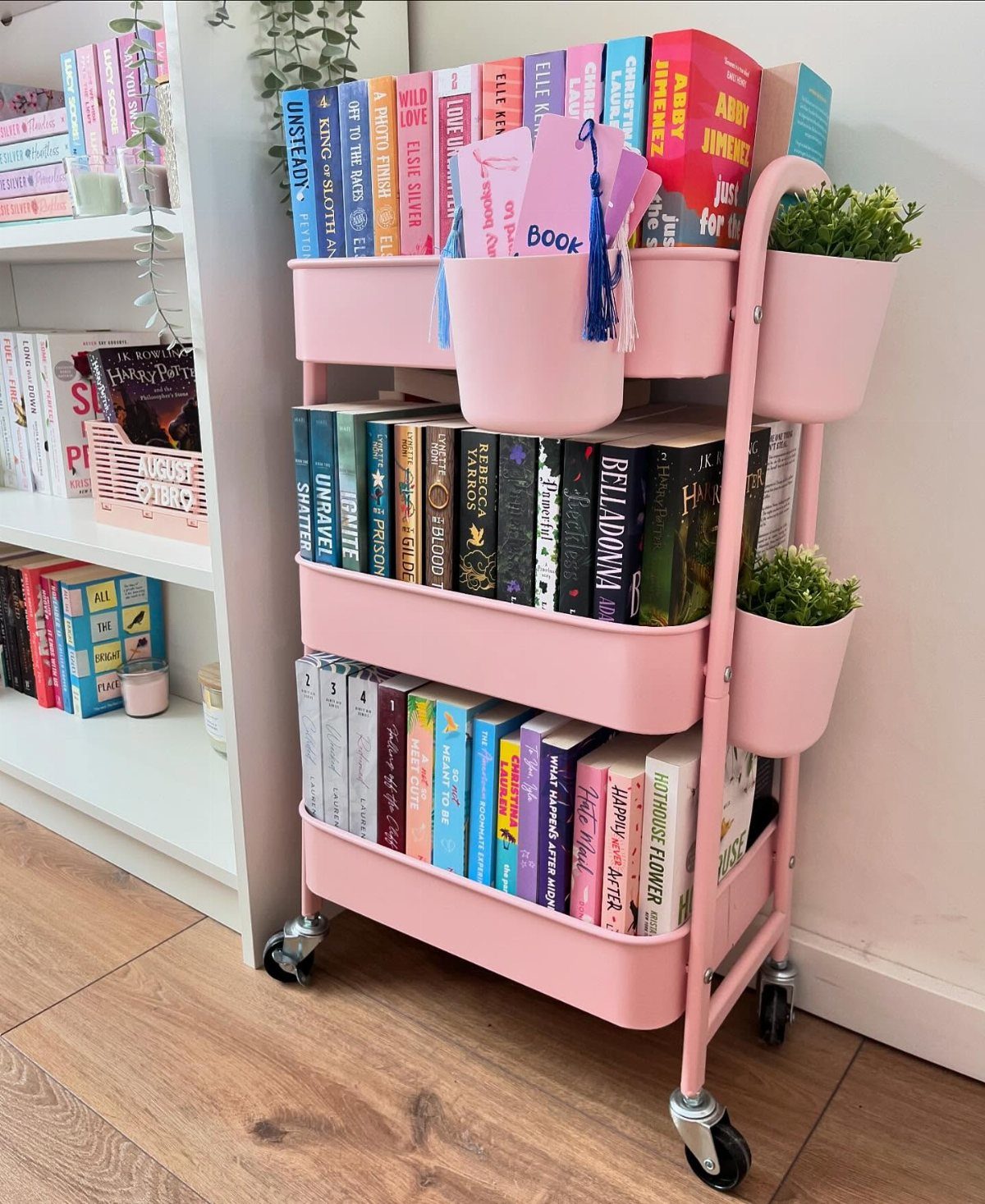 A pink three-tier rolling book cart filled with colorful books and small potted plants.
