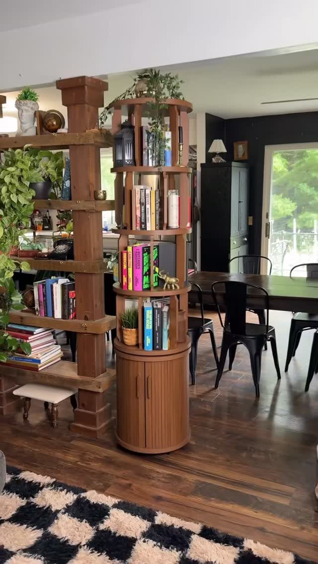 Tall cylindrical wooden bookcase filled with books and plants standing on a dark wood floor.