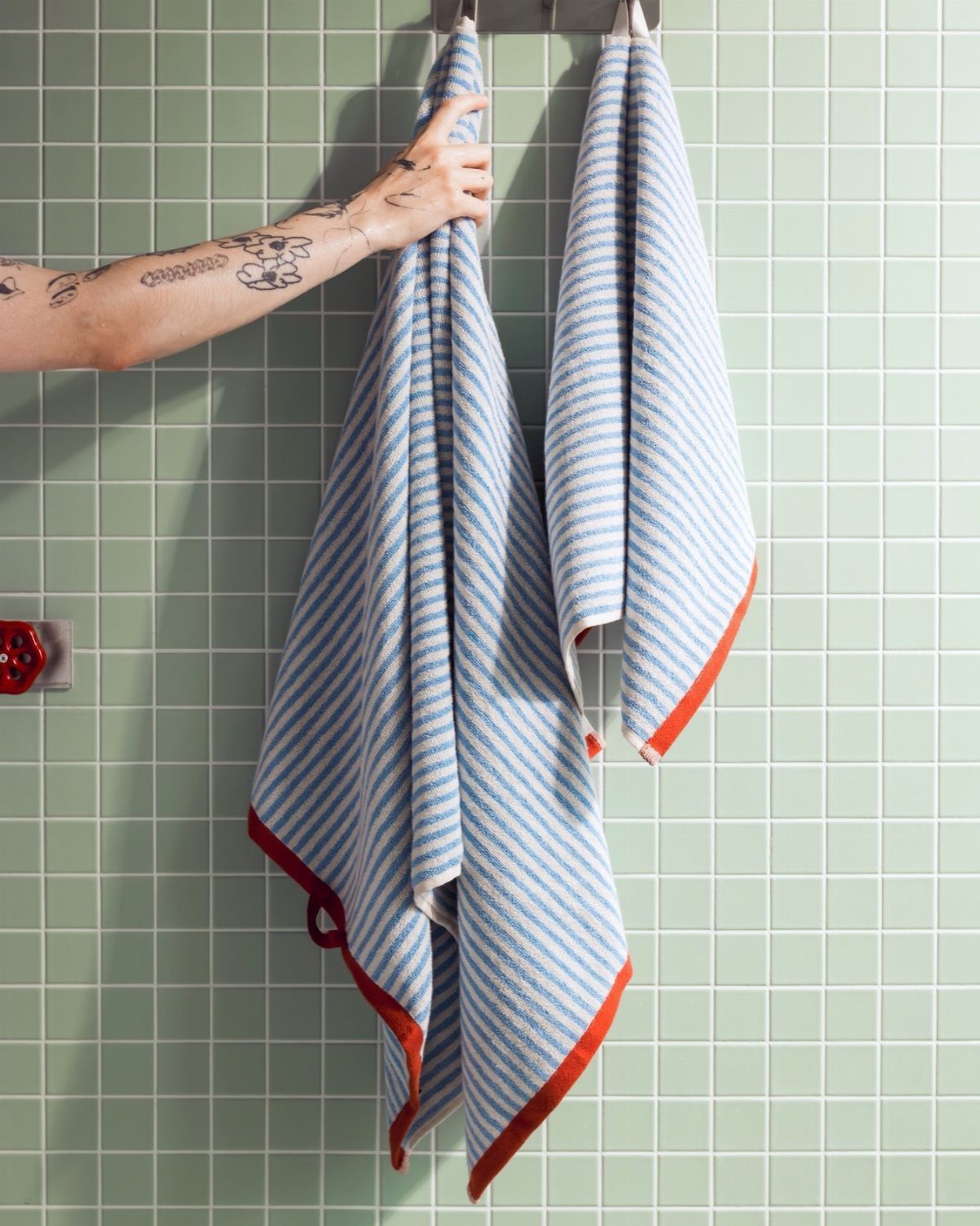 Tattooed arm hanging blue and white striped towels with orange trim on a green tiled wall.
