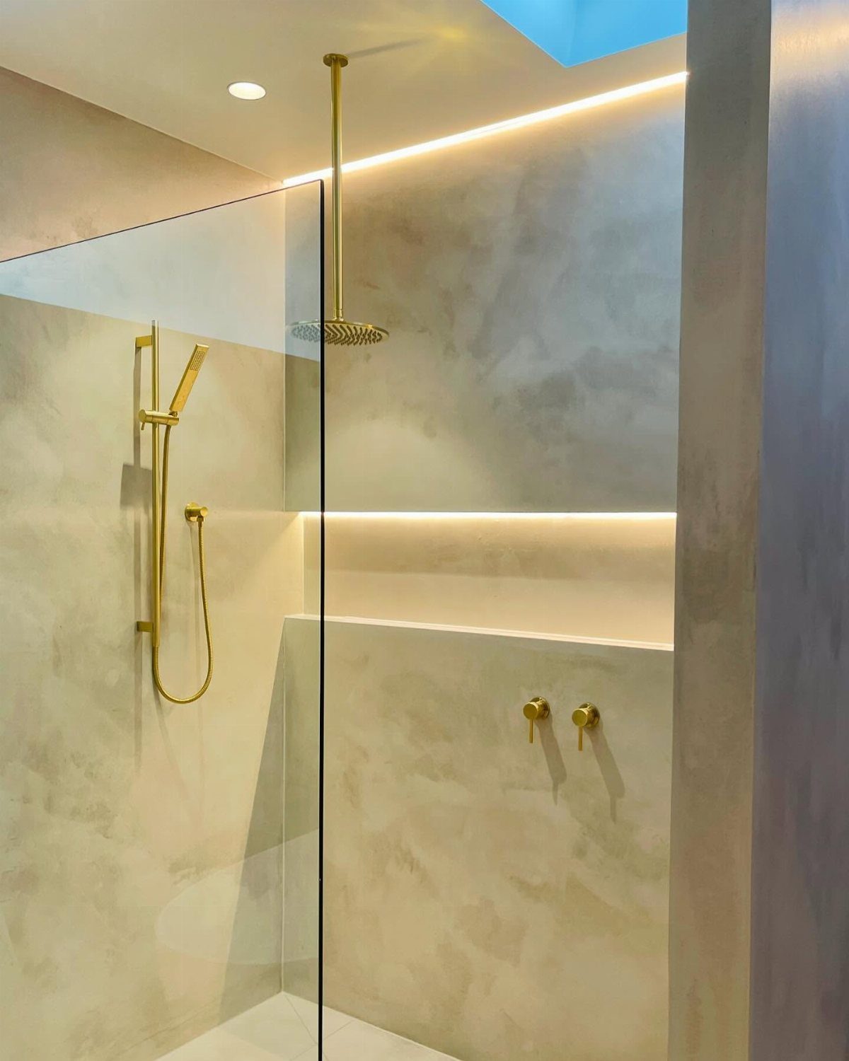 Gold shower fixtures and rain shower head inside a modern beige tiled walk-in shower.