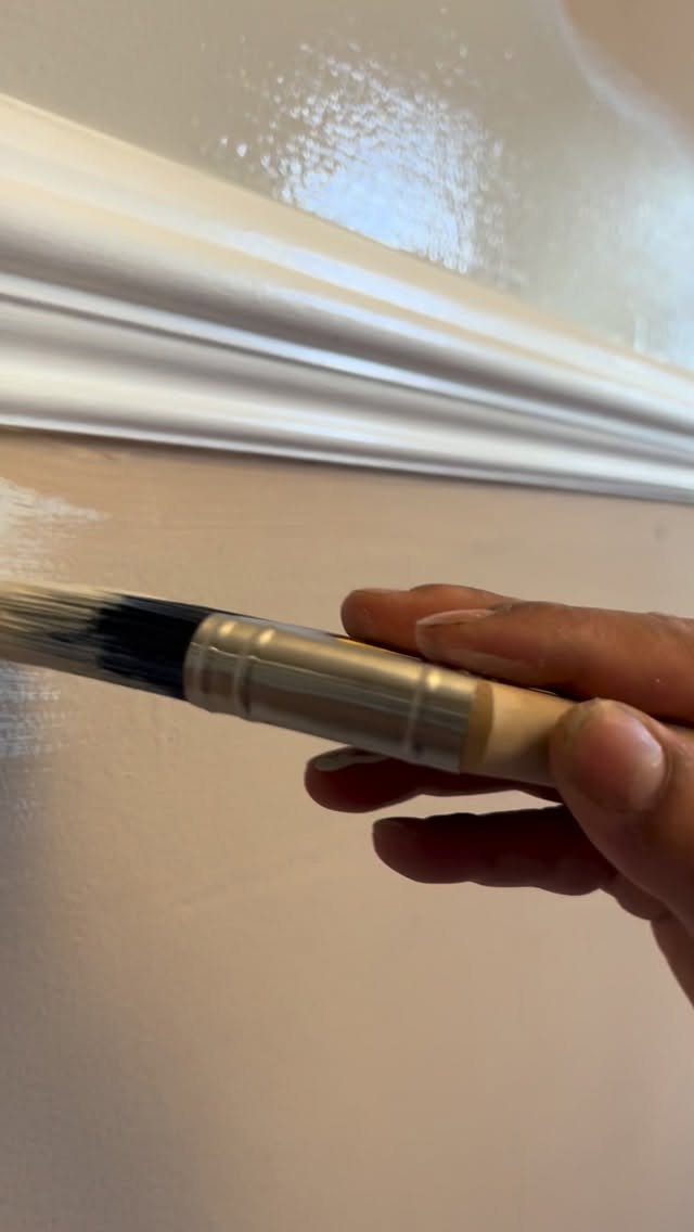 A hand painting a taupe wall beneath white decorative crown molding trim.