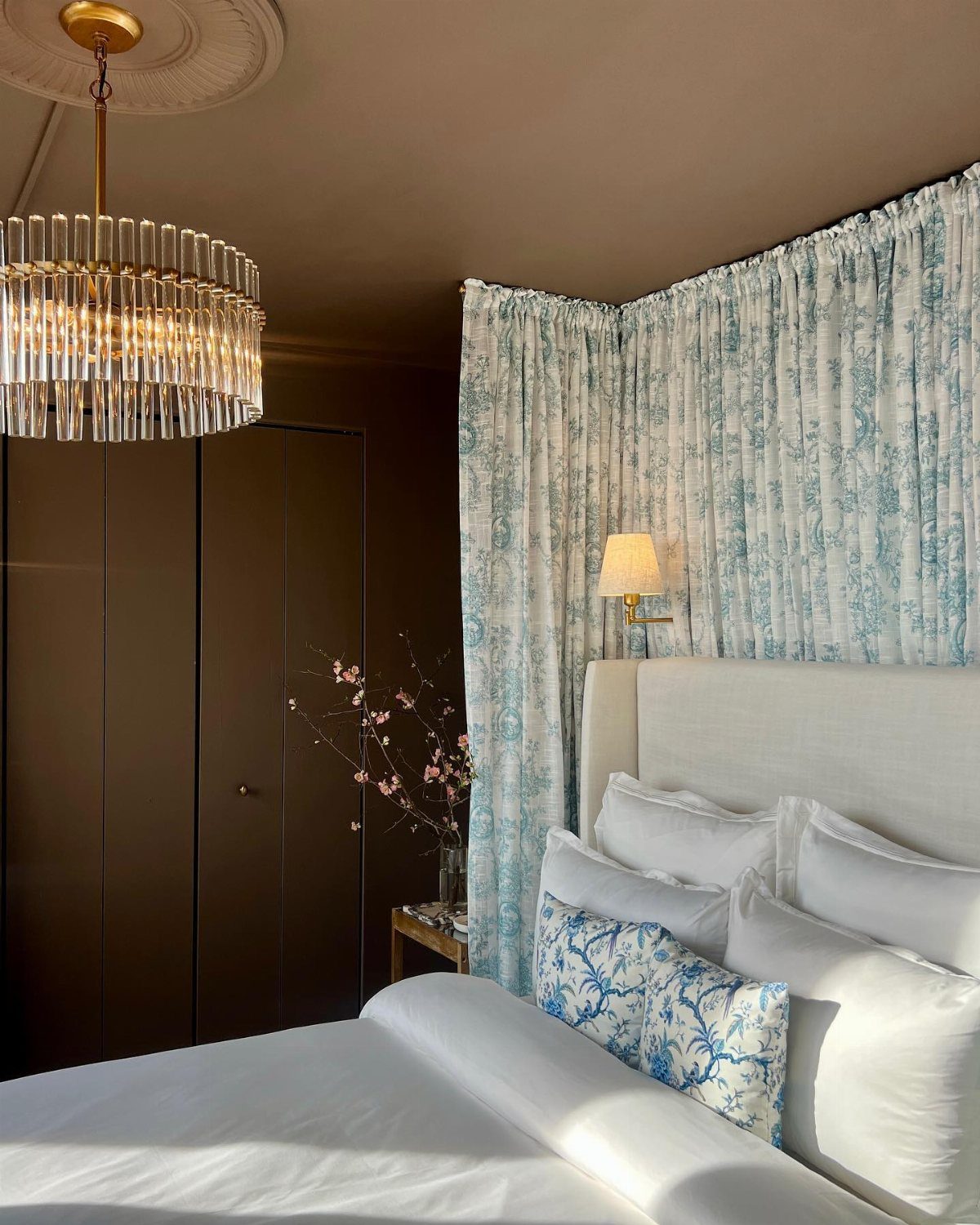 A white bed with blue patterned pillows set against light blue floral curtains and dark walls.