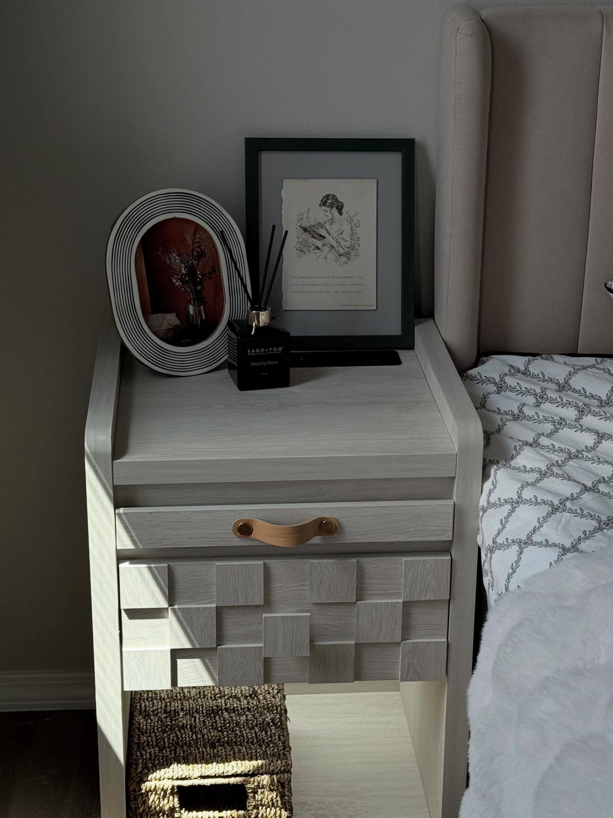 Light grey textured nightstand with a drawer and reed diffuser next to a bed.