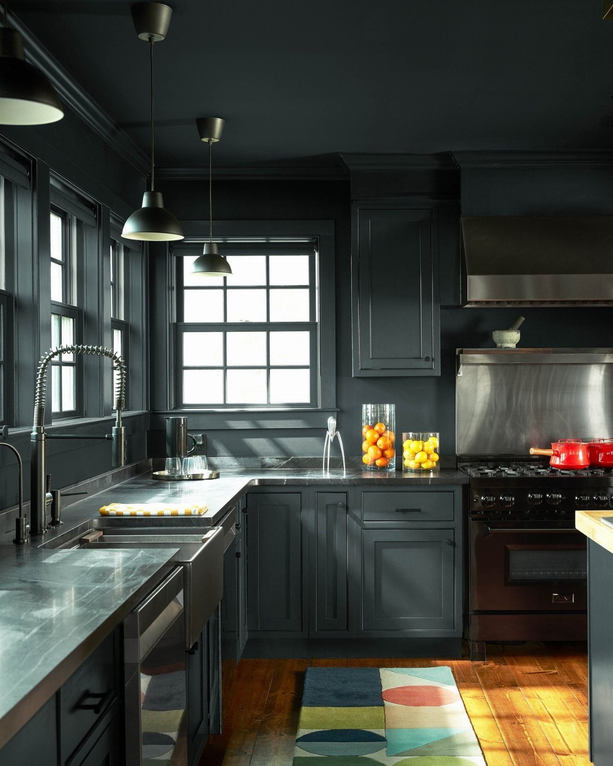 Dark charcoal-green kitchen cabinets with stone countertops and stainless steel appliances near large windows.
