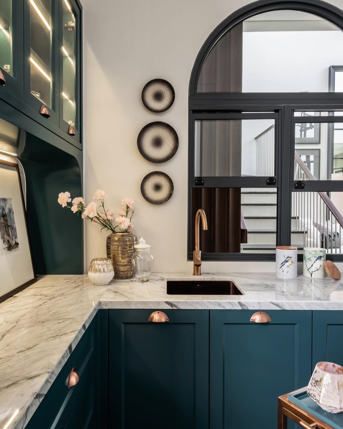 Deep teal green kitchen cabinets with marble countertops and a gold faucet near an arched window.