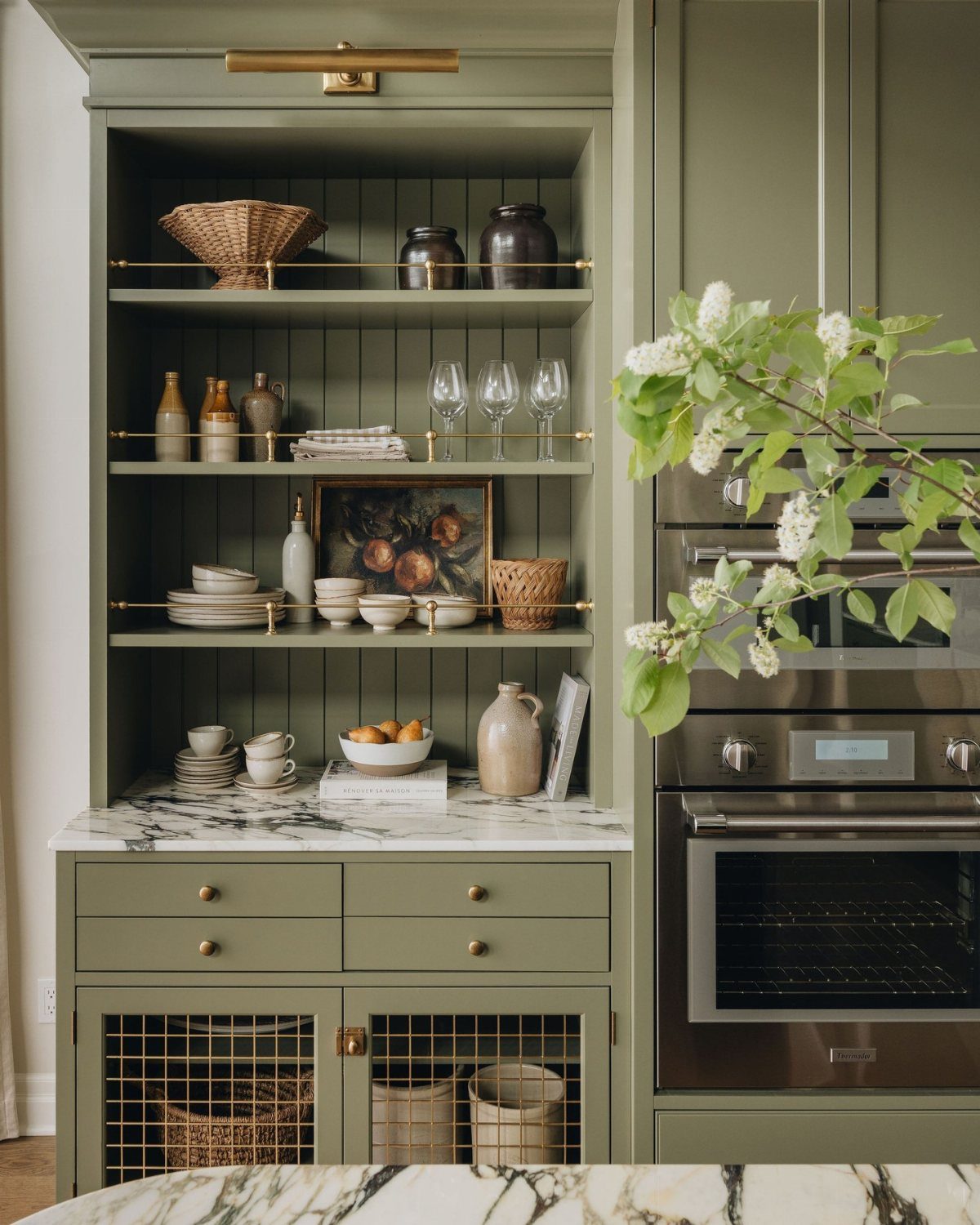 Olive green built-in kitchen hutch with brass accents and marble countertop holding various ceramics.
