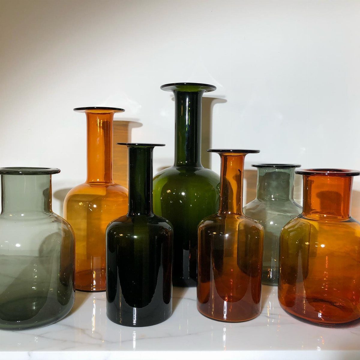 Seven glass vases in amber, green, and gray tones on a marble surface.