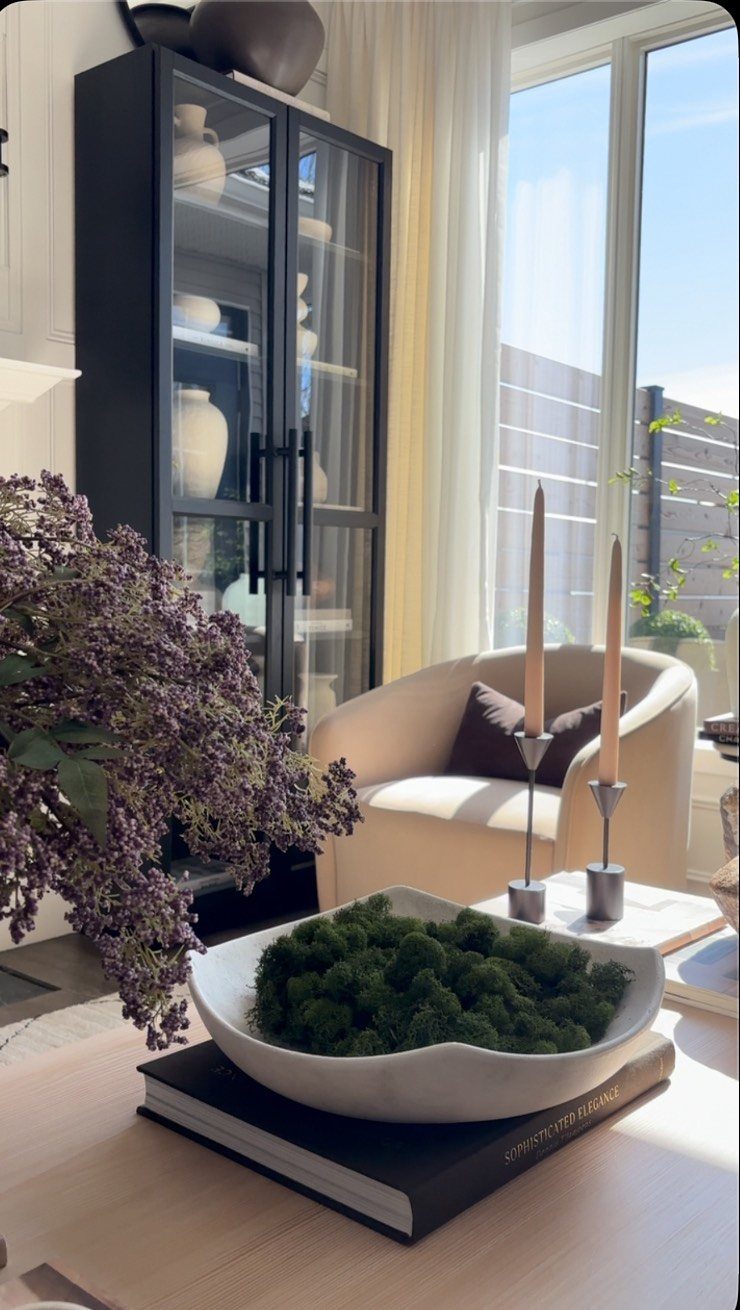 Beige armchair, black display cabinet, purple lilacs, and a moss bowl in a bright living room.