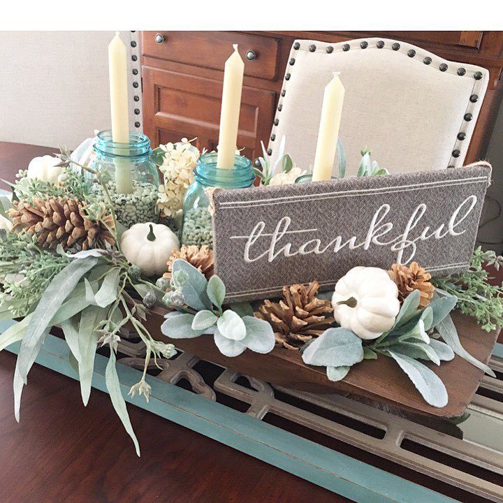 A wooden dough bowl filled with white pumpkins, pinecones, greenery, and candles on a table.