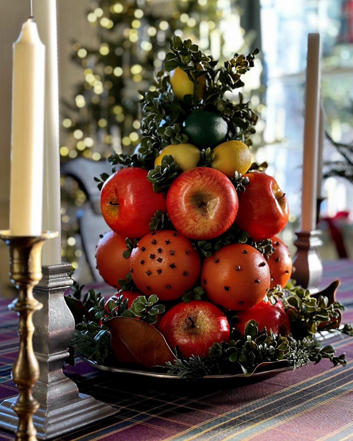 A conical fruit arrangement featuring red apples, oranges, and lemons nestled in green leaves on a table.
