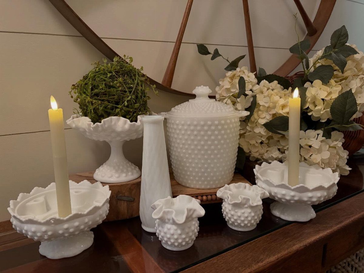 White milk glass containers and bowls with lit taper candles and white hydrangeas on a wooden table.