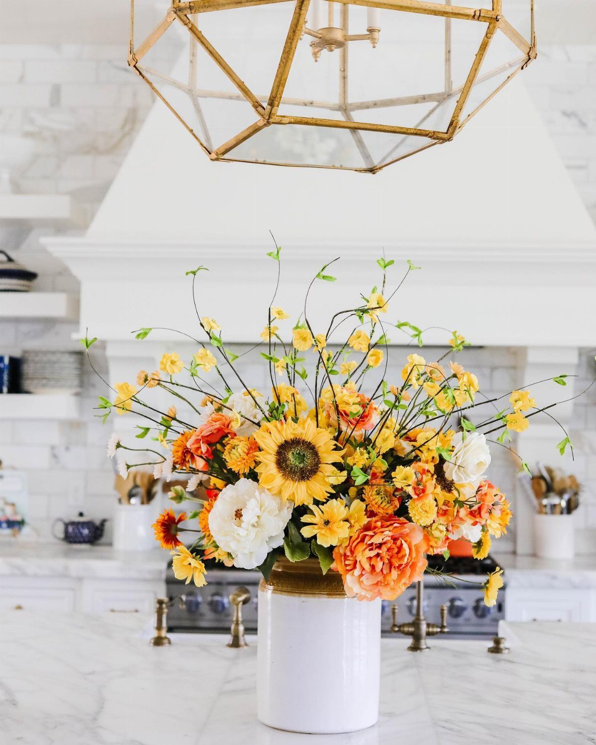 Sunflowers, orange roses, and white peonies in a white vase on a marble counter.