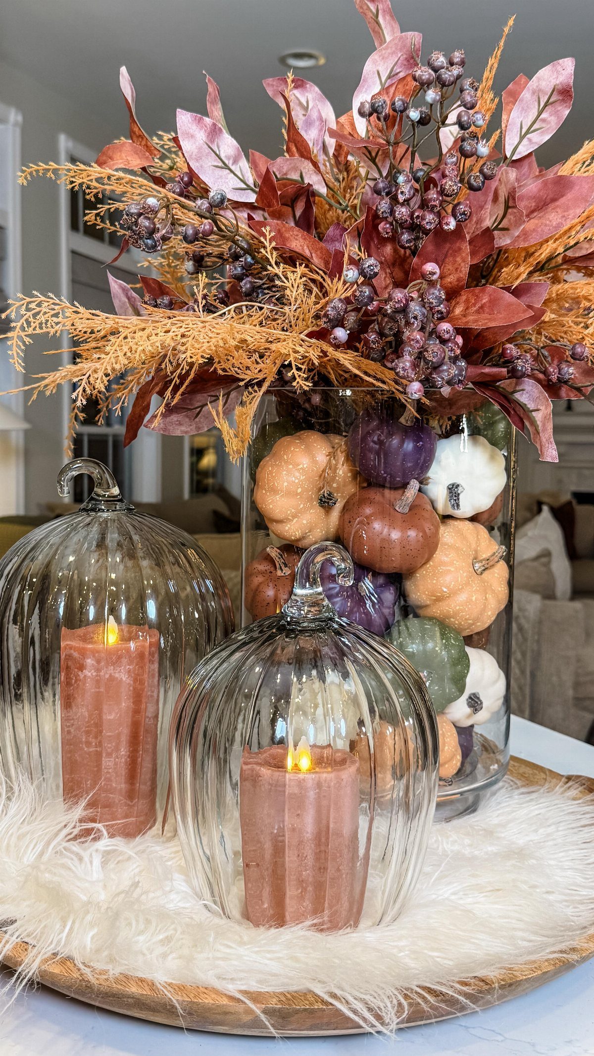 Autumn centerpiece on a wooden tray featuring glass pumpkin candleholders and a vase of pumpkins and fall foliage.