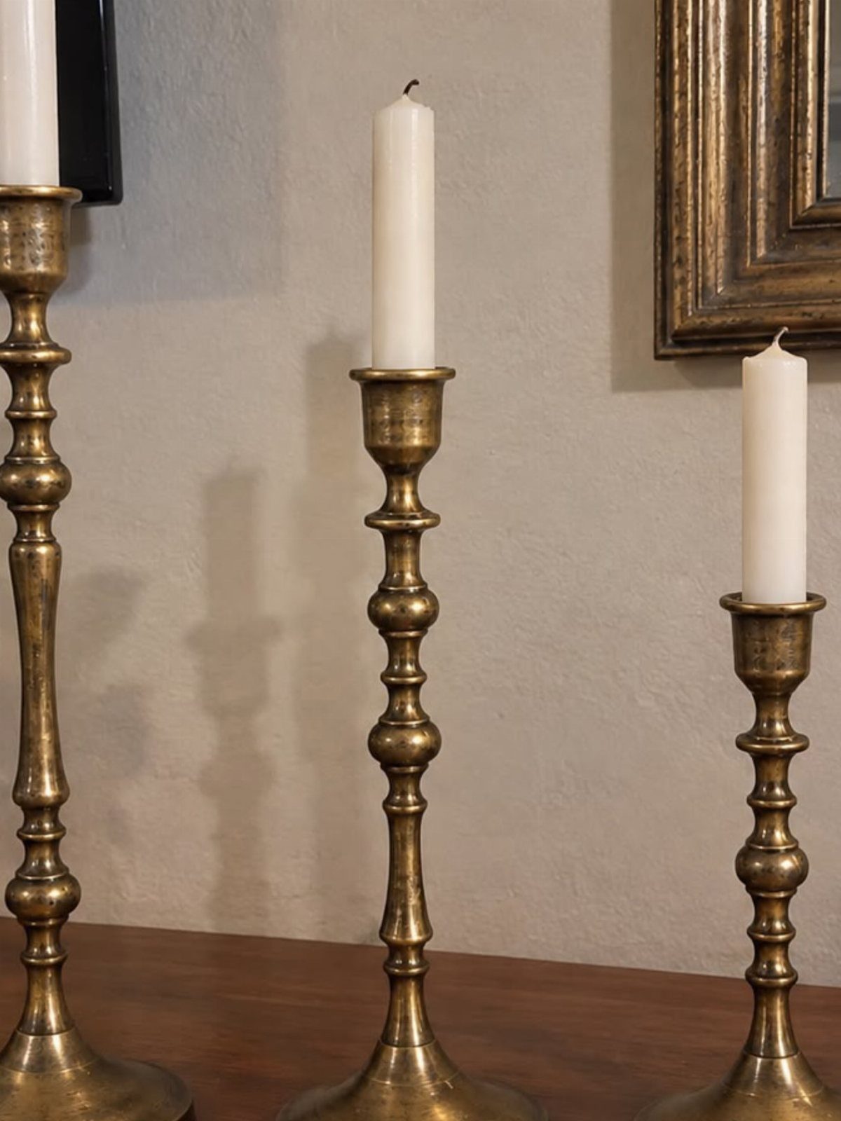 Three mismatched brass candlesticks with white taper candles on a wooden table.