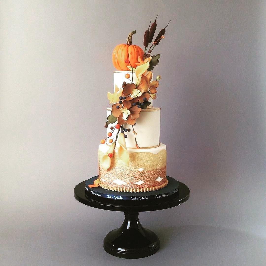 A tiered white cake decorated with a pumpkin and autumn leaves on a black cake stand.