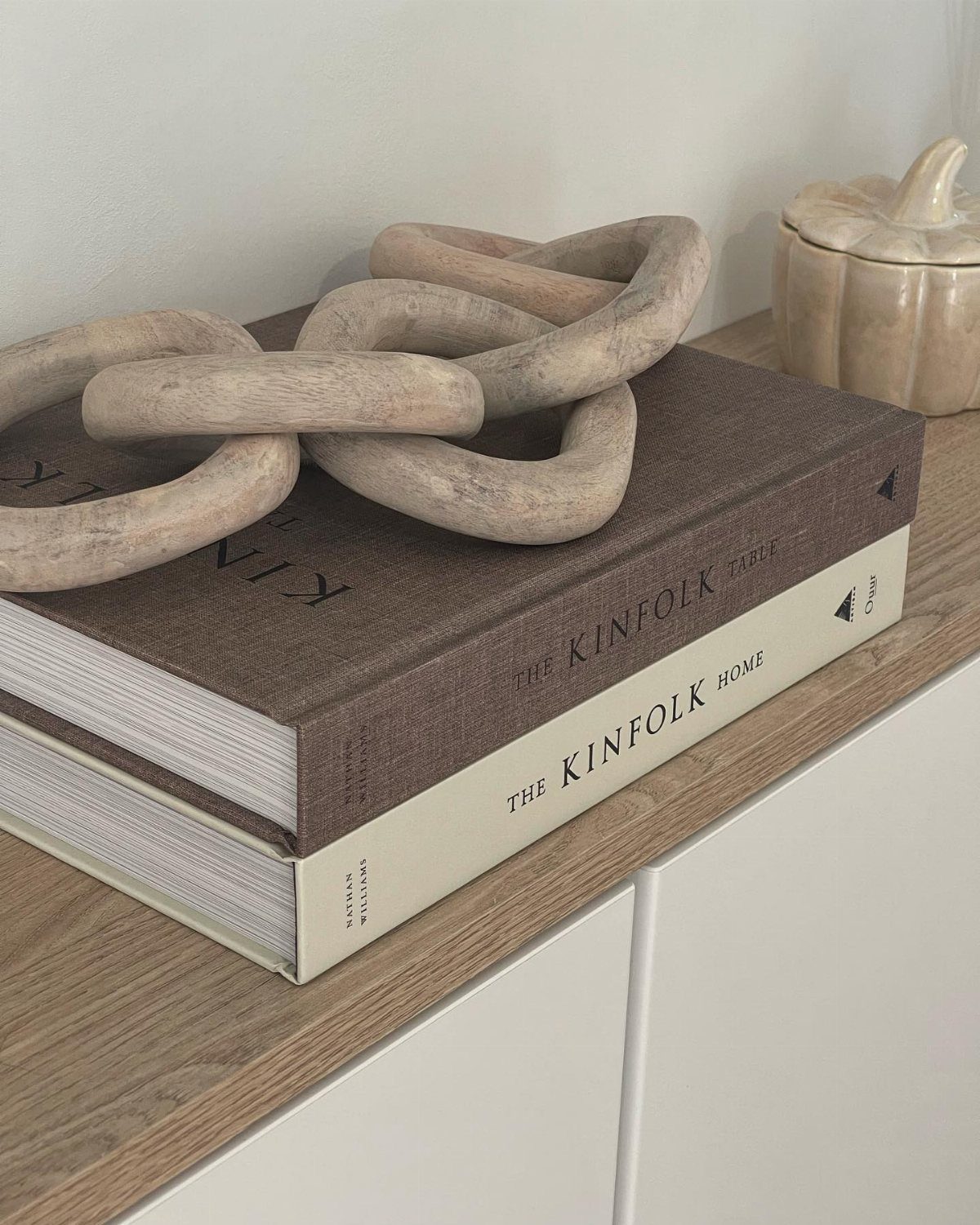 Stacked hardcover books with a large wooden chain sculpture on a light wood shelf.