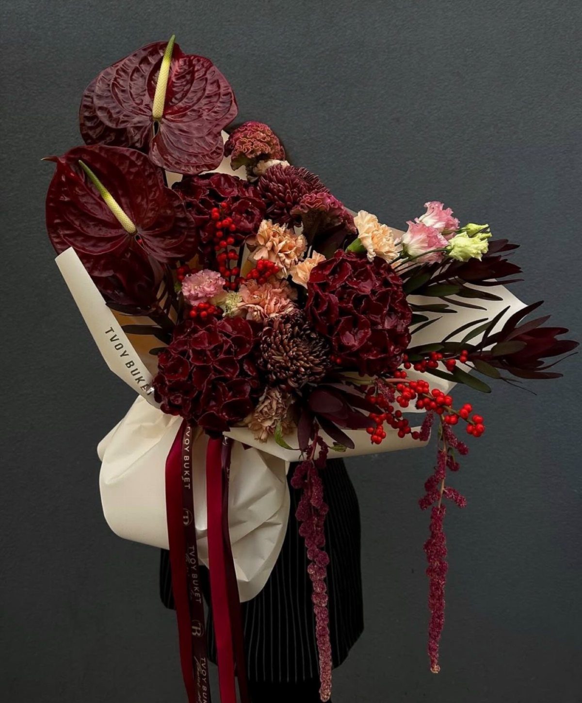 A large bouquet of deep burgundy anthuriums, carnations, and red berries wrapped in white paper.
