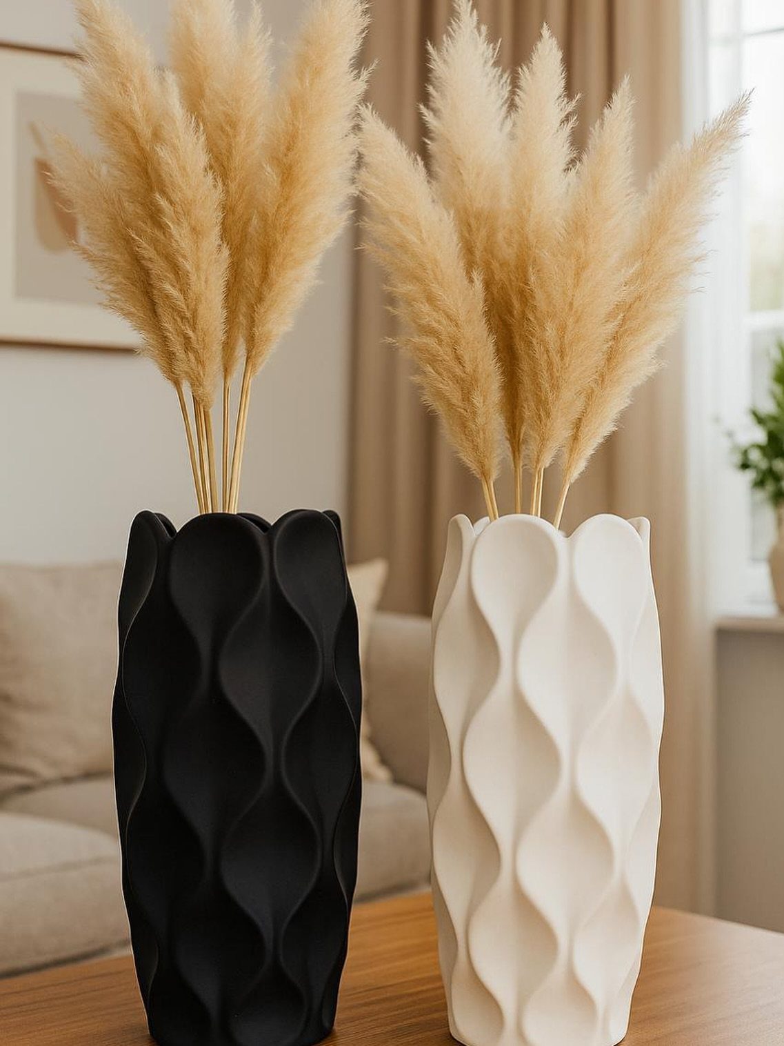 Two textured sculptural vases, one black and one cream, holding dried pampas grass on a wooden table.