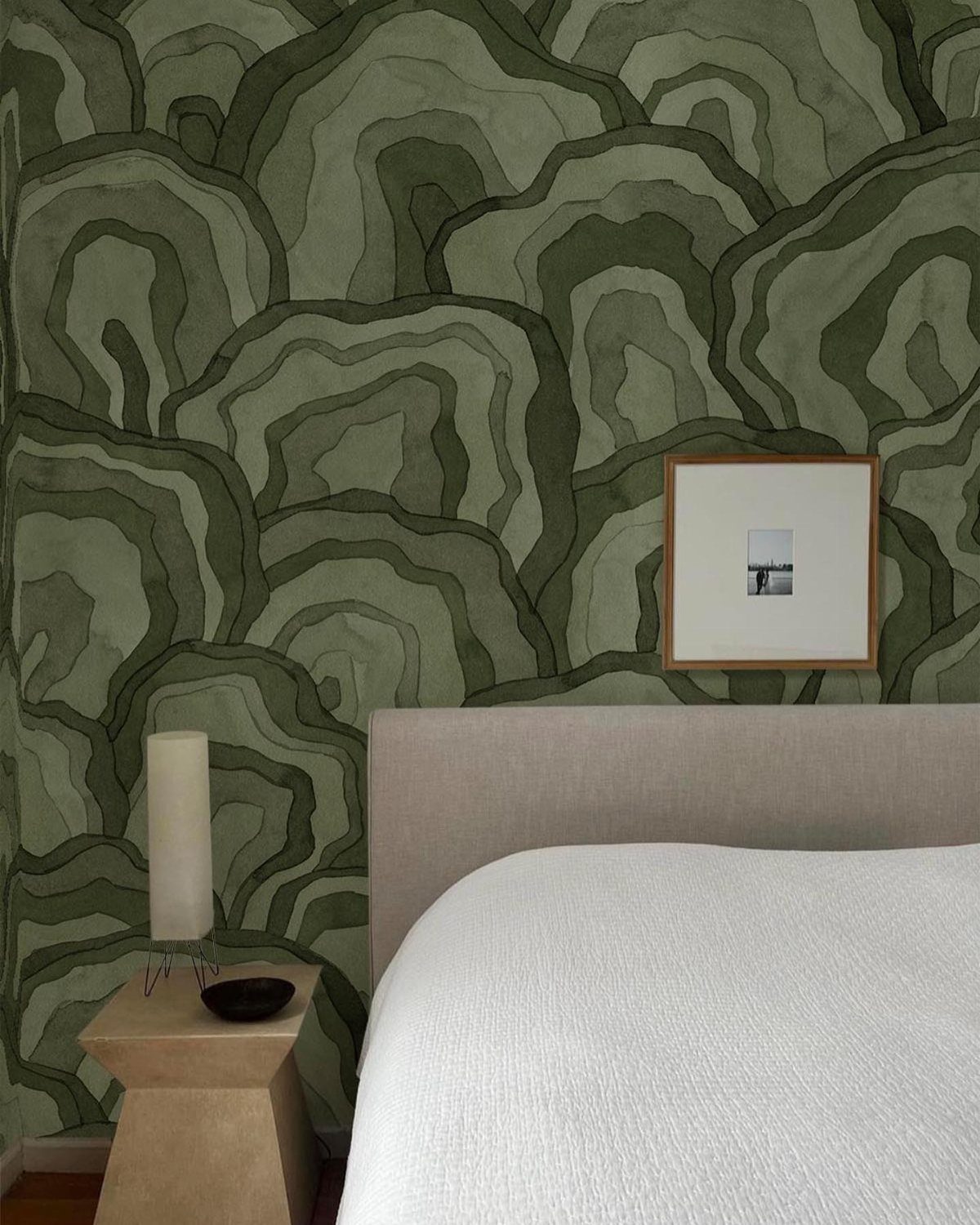 Green patterned wallpaper behind a bed with white linens and a wooden bedside table.