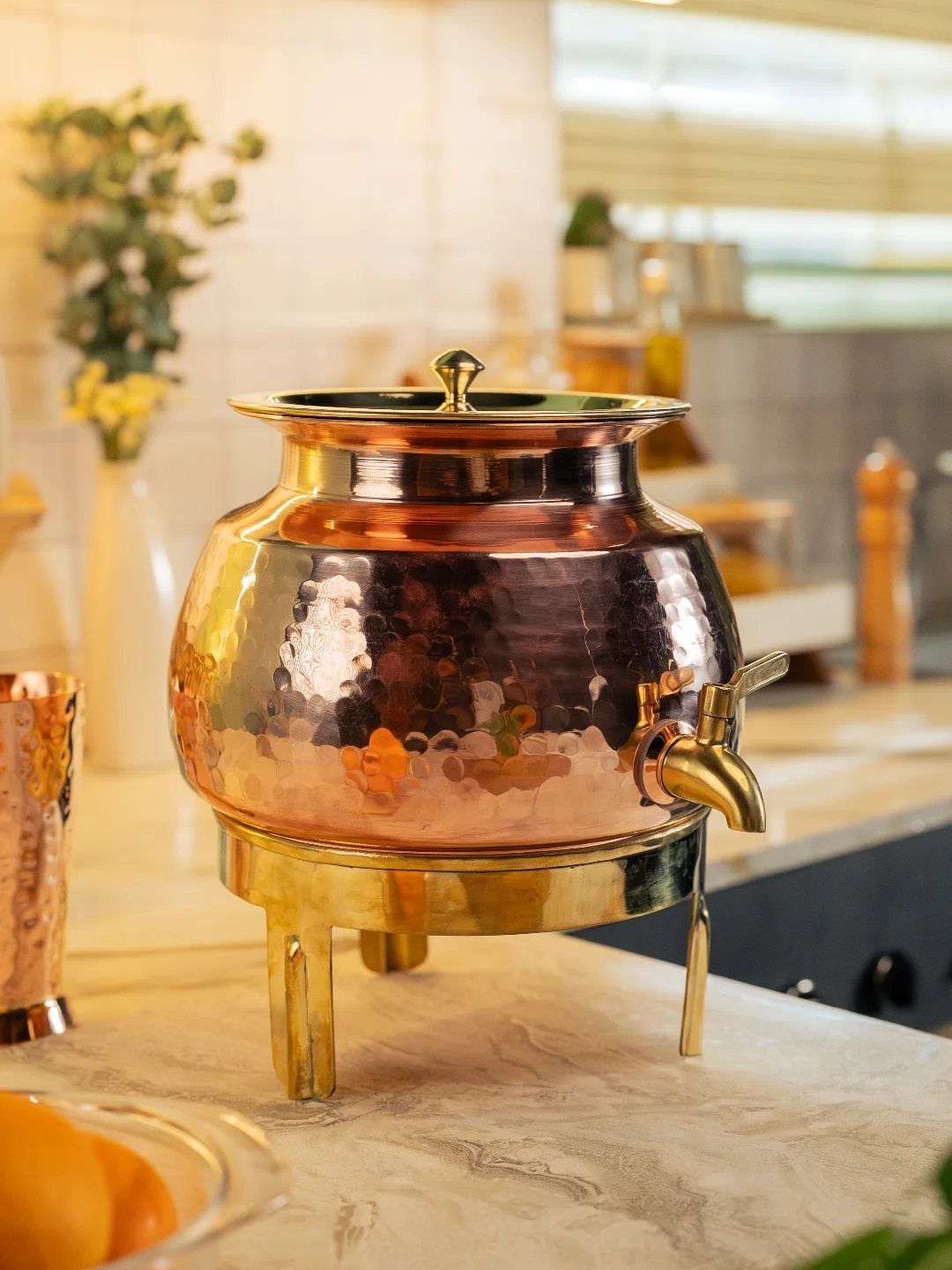A hammered copper beverage dispenser with a gold base sits on a marble kitchen countertop.