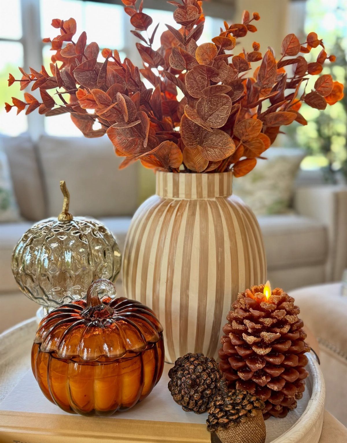 Decorative tray featuring glass pumpkins, a striped vase with orange leaves, and a pinecone candle.