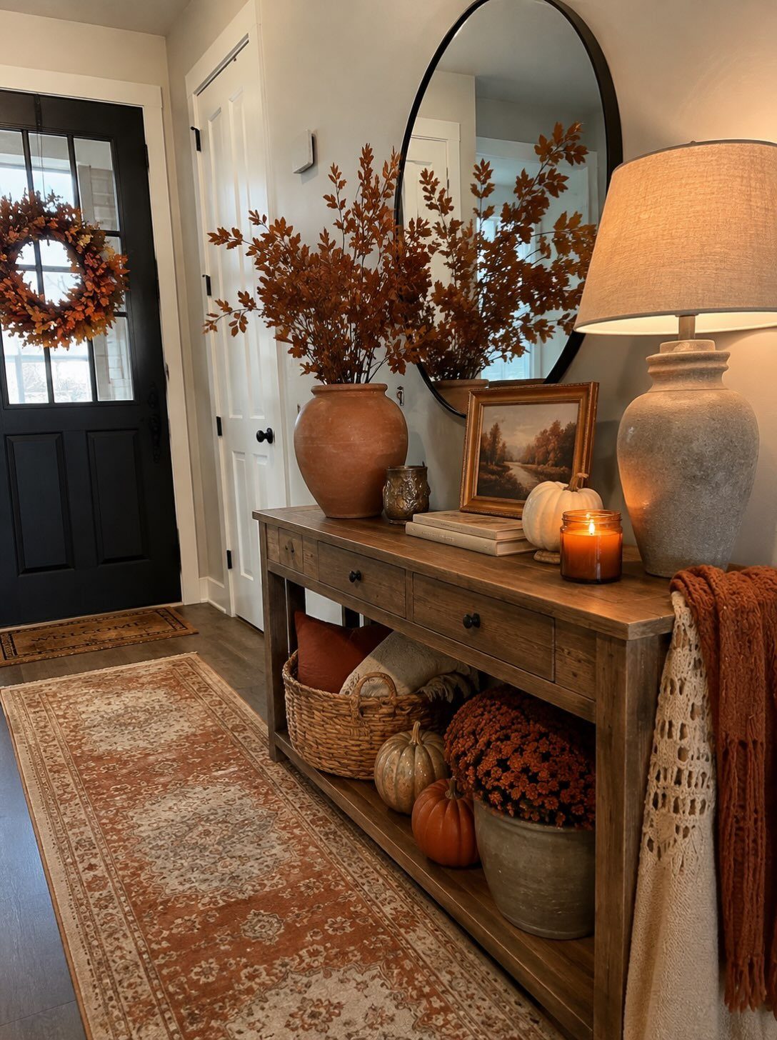Wooden entryway console table decorated with autumn leaves, pumpkins, a candle, and a lamp.