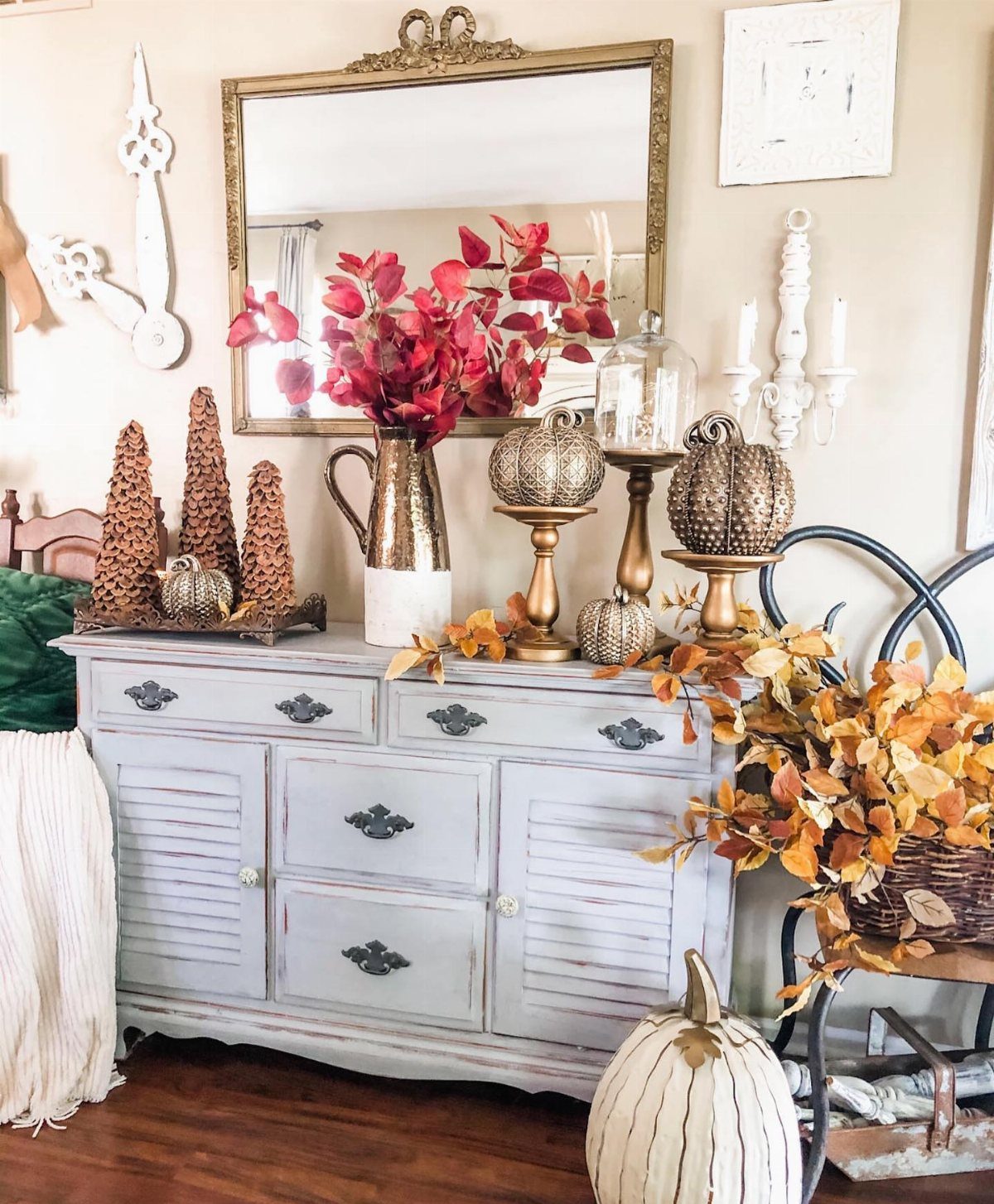 White sideboard decorated with red flowers, metallic pumpkins, and autumn foliage under a gold mirror.