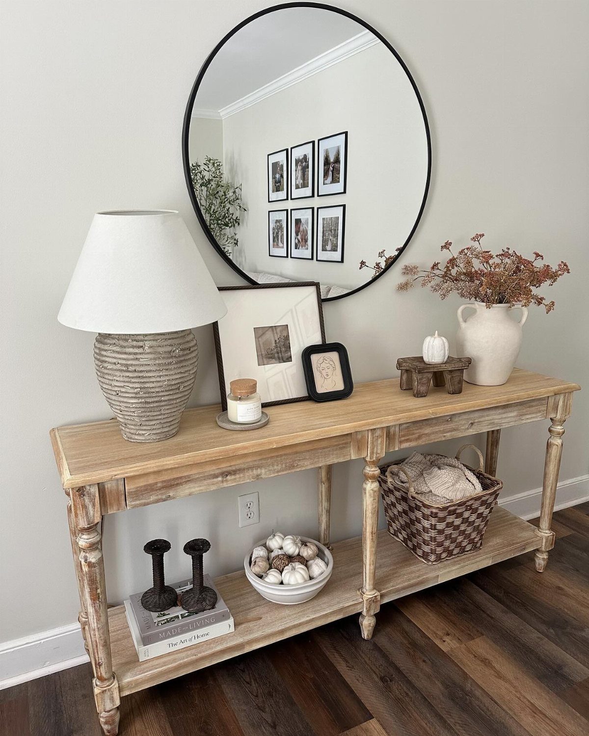Wooden console table decorated with a round mirror, lamp, and dried autumn branches.