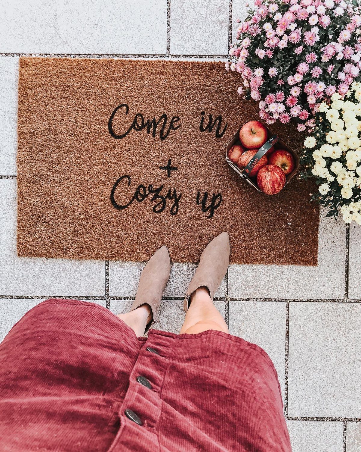 Brown welcome mat with text on a tiled floor next to autumn flowers and apples.