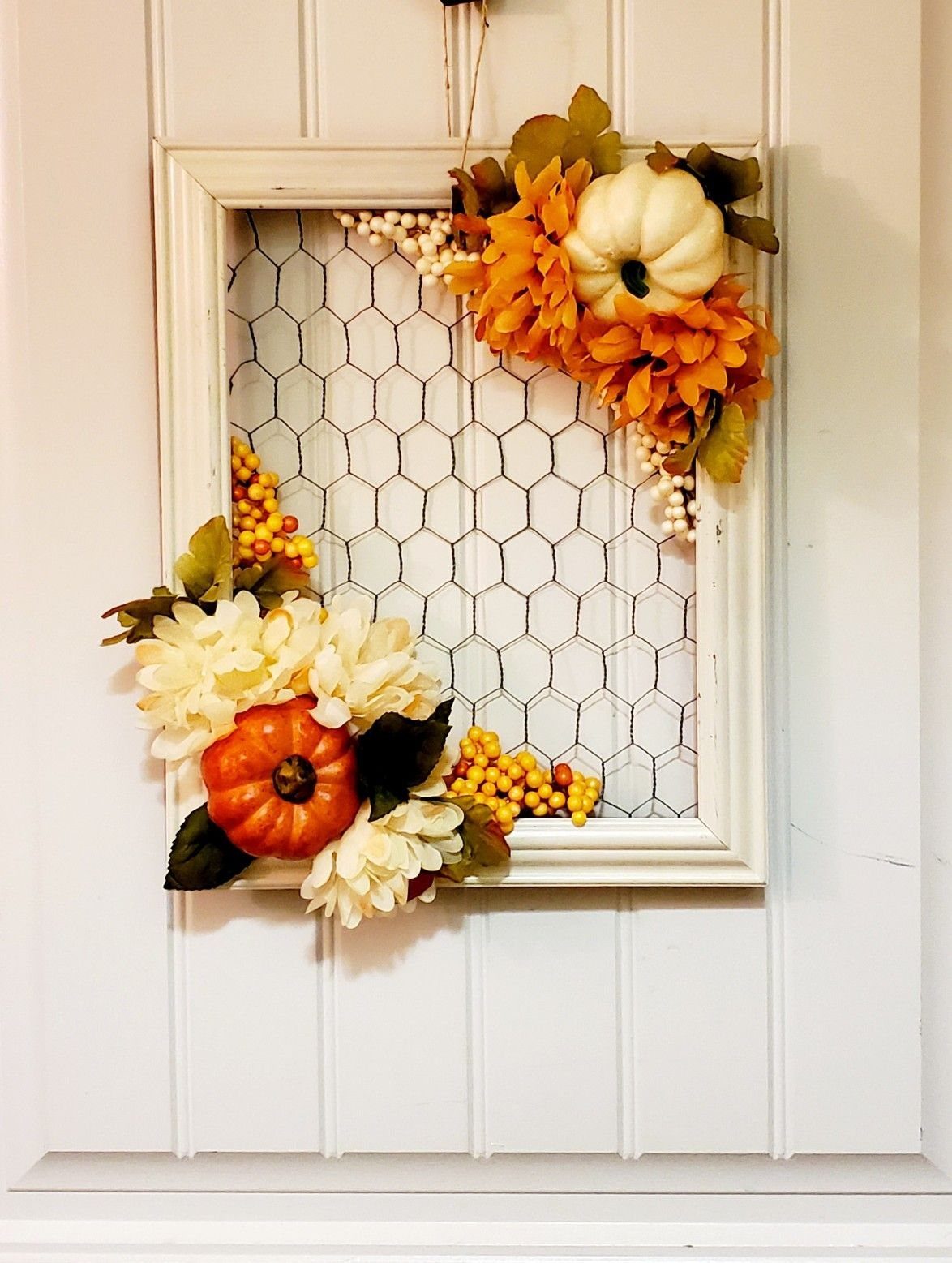 White decorative picture frame with chicken wire and autumn floral accents hanging on a white door.