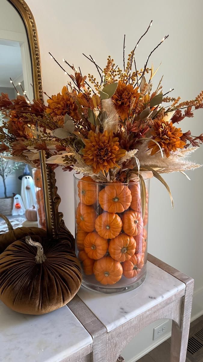 Clear glass vase filled with small pumpkins and autumn floral arrangement on a marble table.