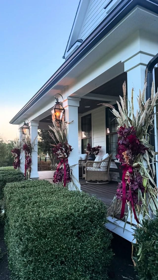 Decorated cornstalks with burgundy flowers attached to white porch columns of a house.