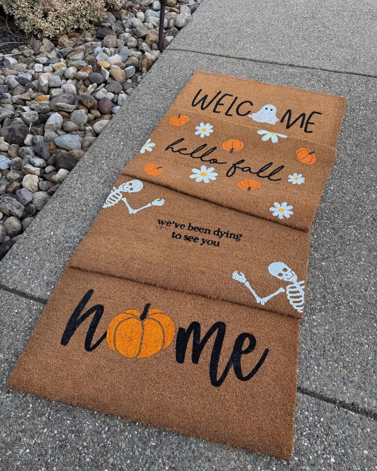 Three decorative coir doormats with fall and Halloween themes laid on a concrete walkway.