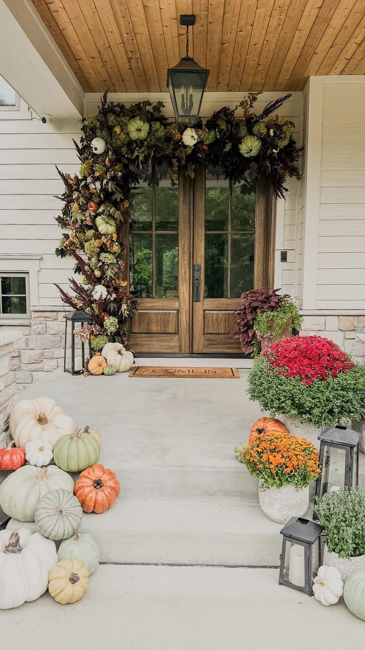 Autumn porch decor with pumpkins, potted flowers, and a leafy garland surrounding wooden front doors.