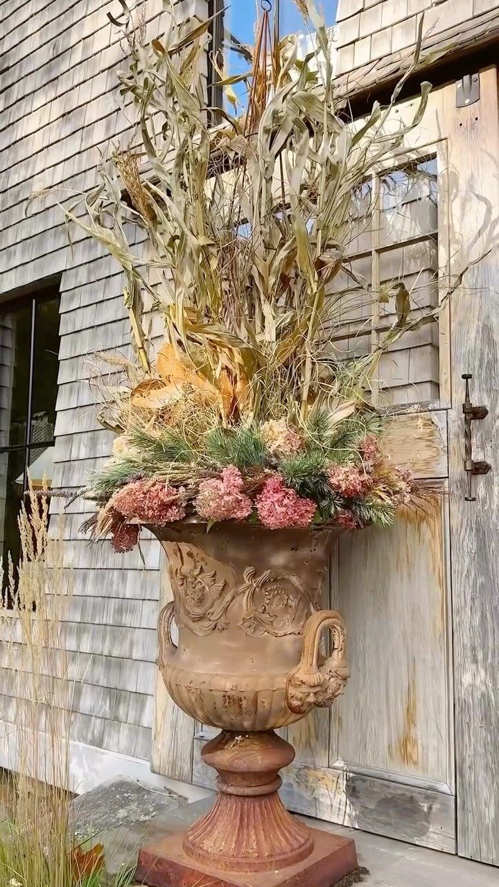 Dried cornstalks and pink hydrangeas arranged in a large, decorative terracotta urn on a porch.