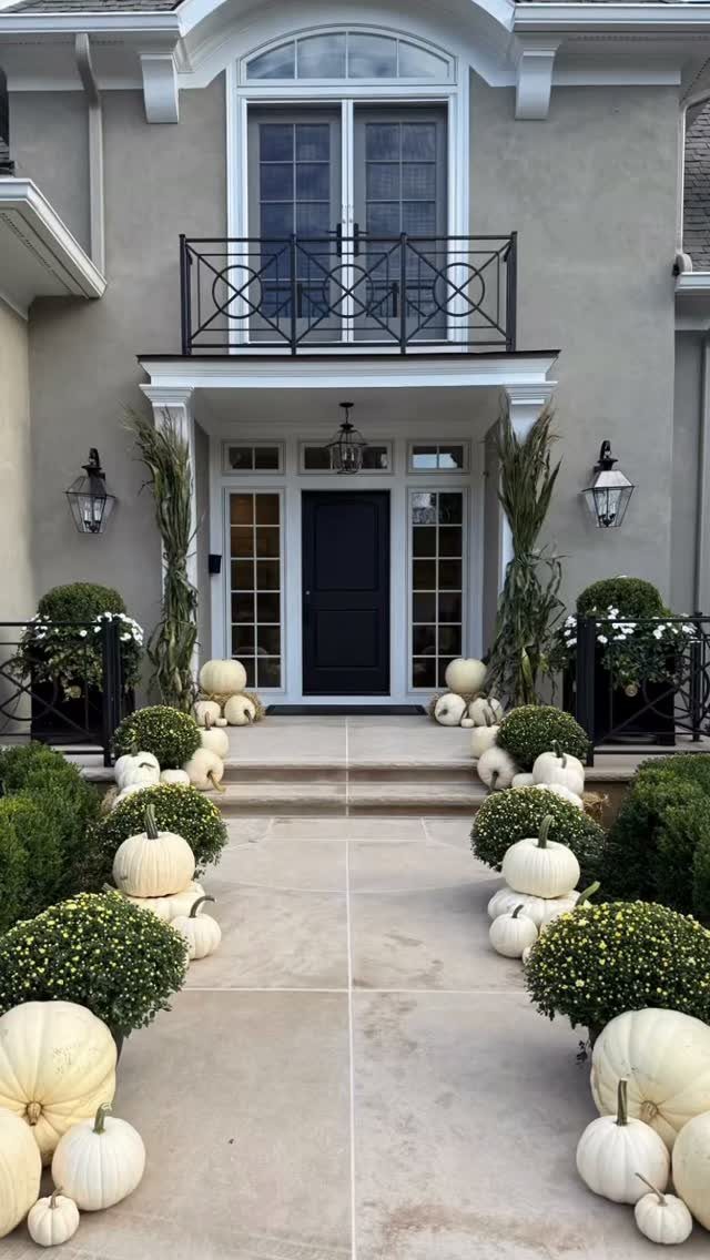 White pumpkins and green shrubs line a stone walkway leading to a house entrance decorated for fall.