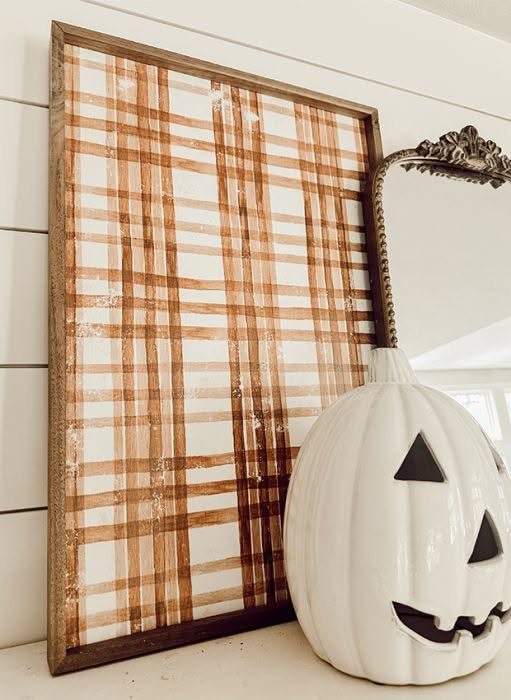 Framed plaid art leaning against a white wall next to a white ceramic pumpkin.