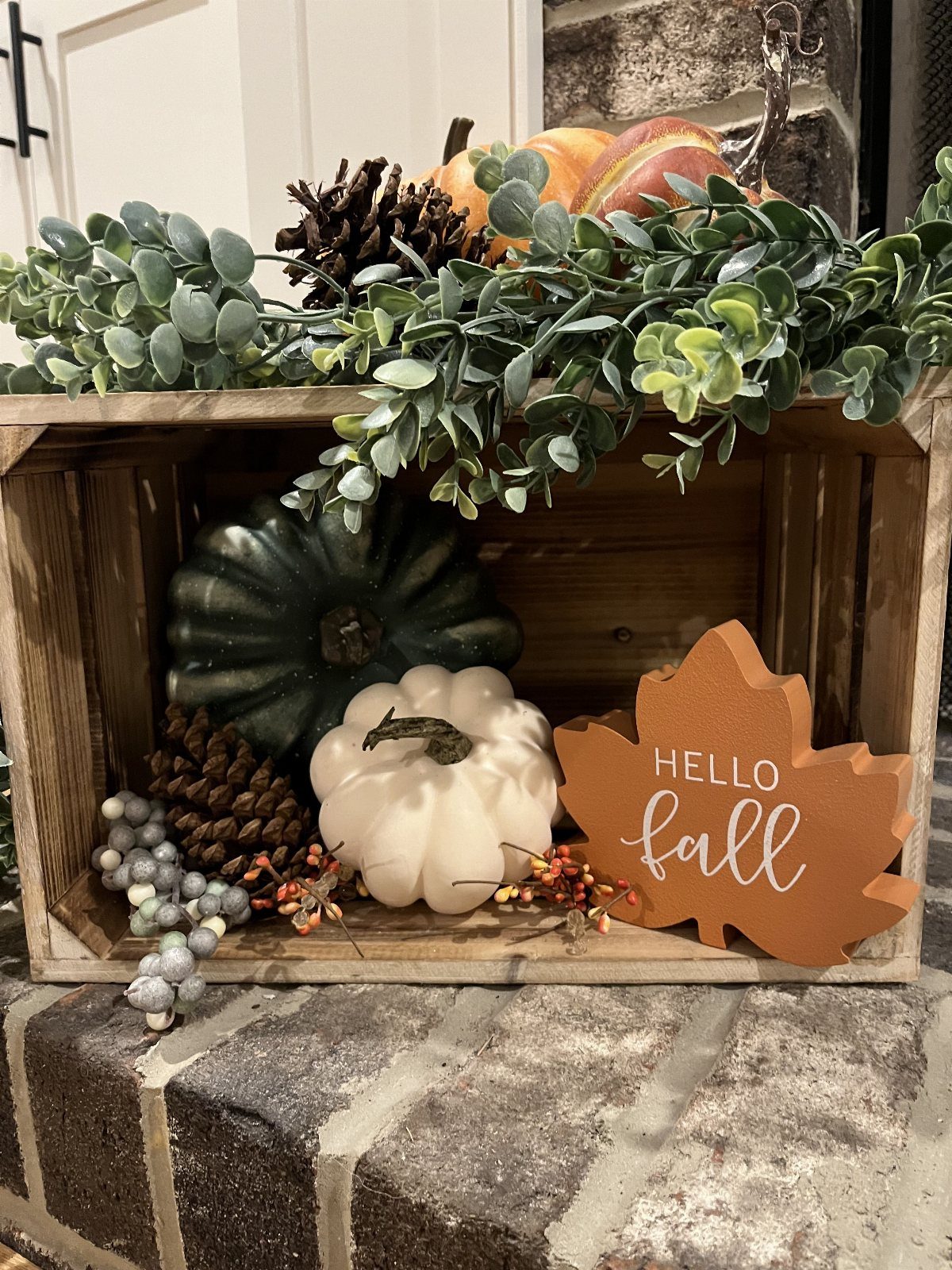 Wooden crate filled with pumpkins, pinecones, greenery, and a fall leaf sign on a brick surface.