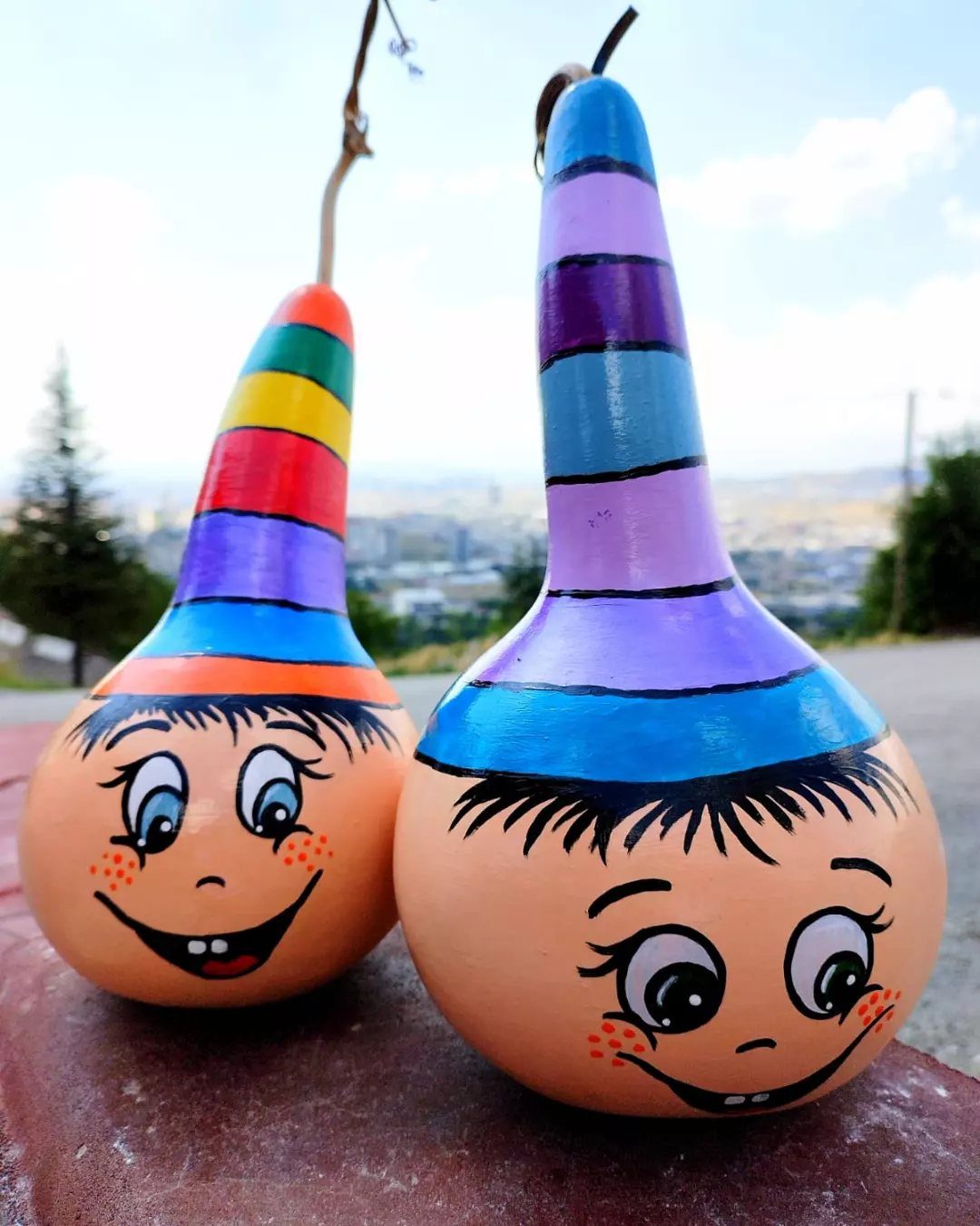 Two painted gourds with colorful striped hats and cartoon faces sitting outdoors.