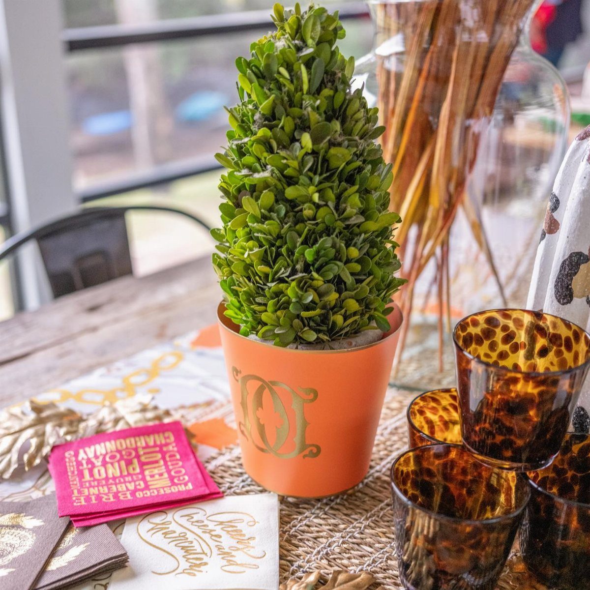 Small green boxwood plant in an orange monogrammed pot on a decorated wooden table.