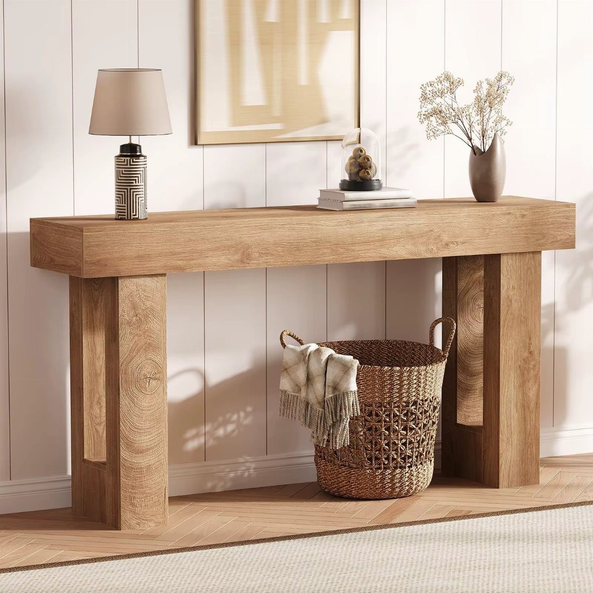 A light wood console table decorated with a lamp, books, and a vase of dried flowers.