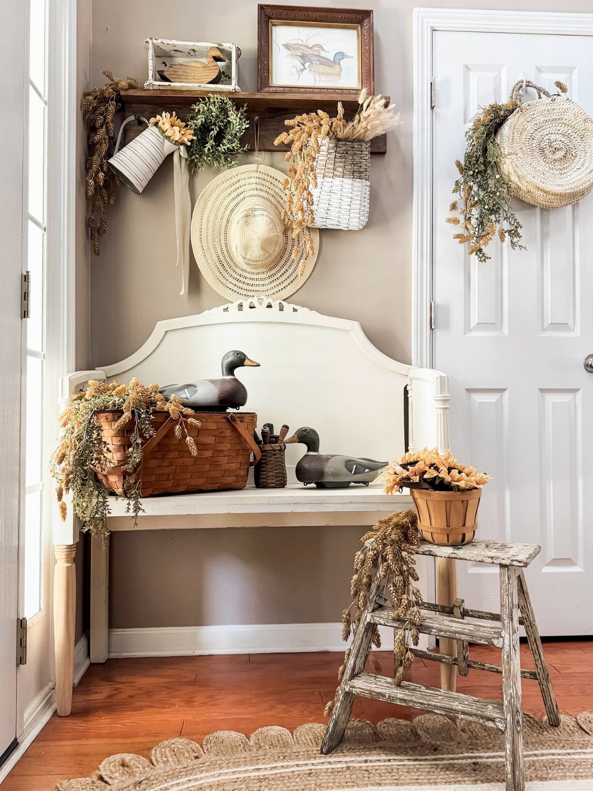 White wooden bench with dried flowers, duck figurines, and a straw hat in a rustic entryway.