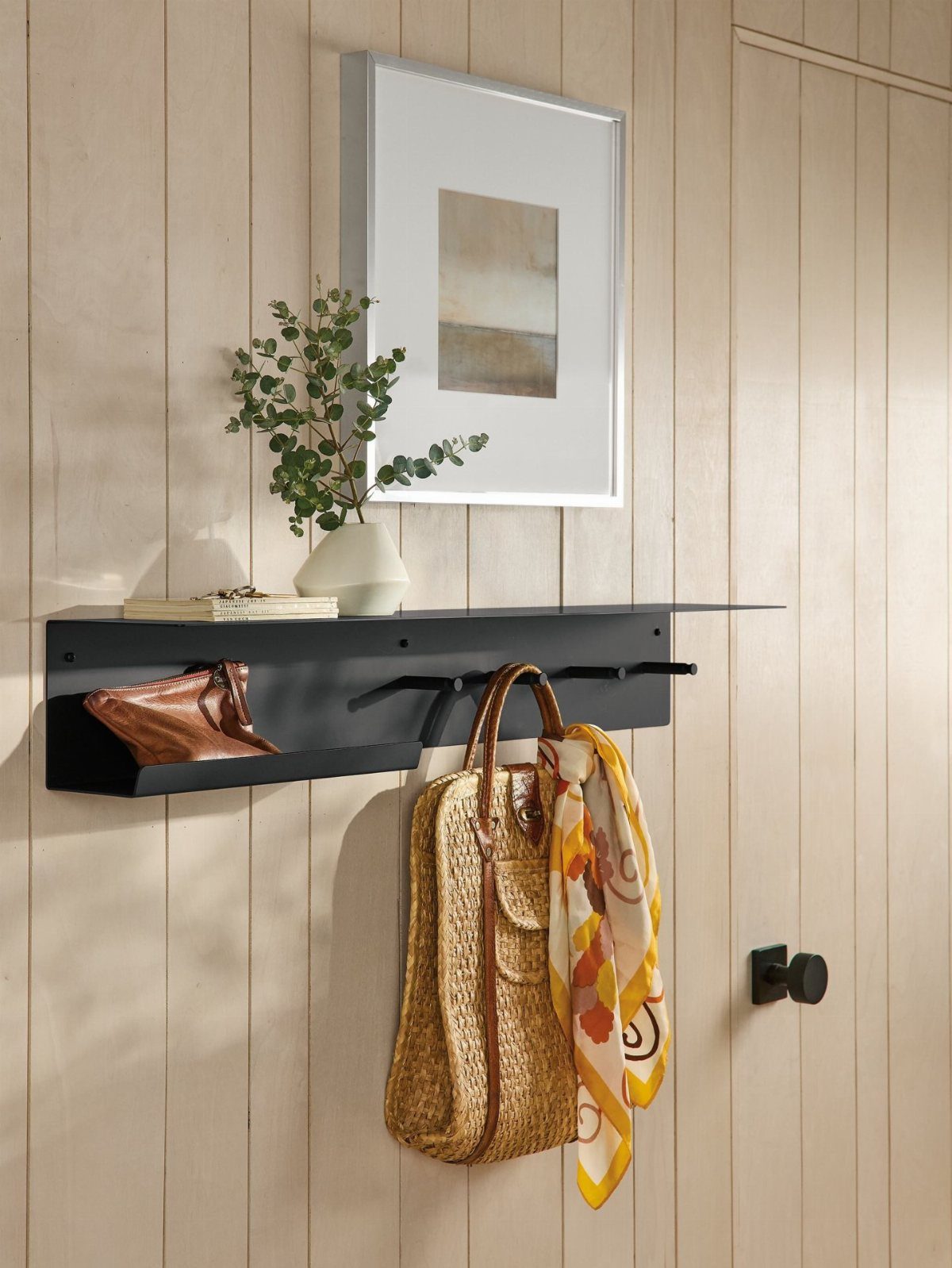 Black wall-mounted shelf with hooks holding a straw bag and a colorful scarf.