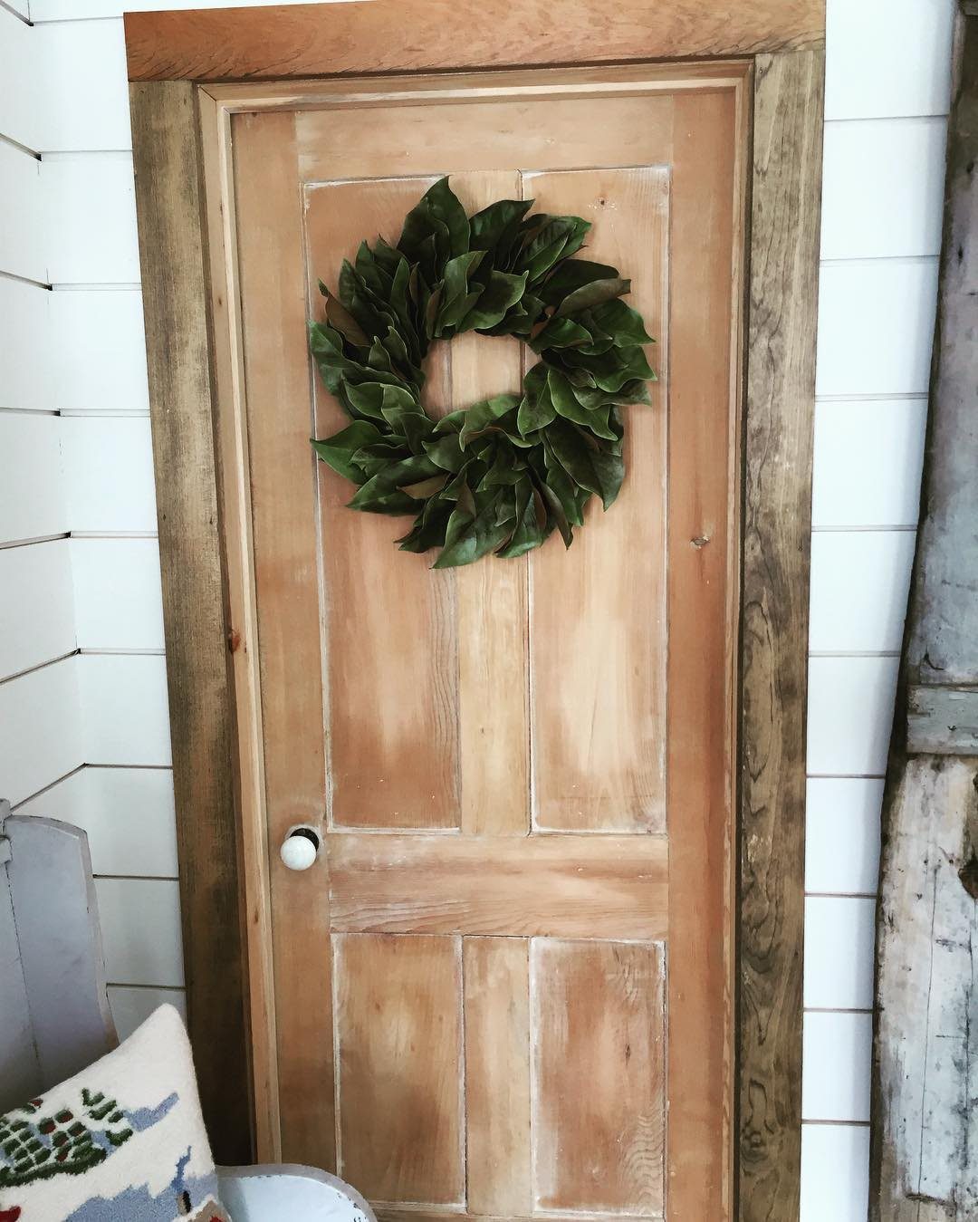 A green leafy wreath hanging on a light wood paneled door with white shiplap walls.