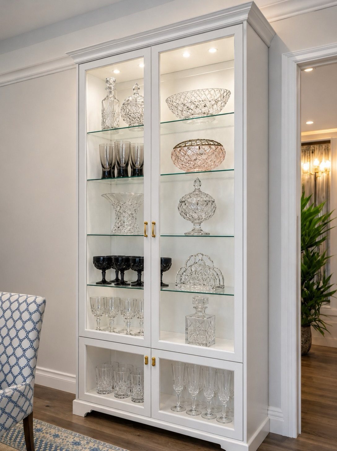 White glass-front display cabinet with beadboard backing containing various crystal glassware and decanters.
