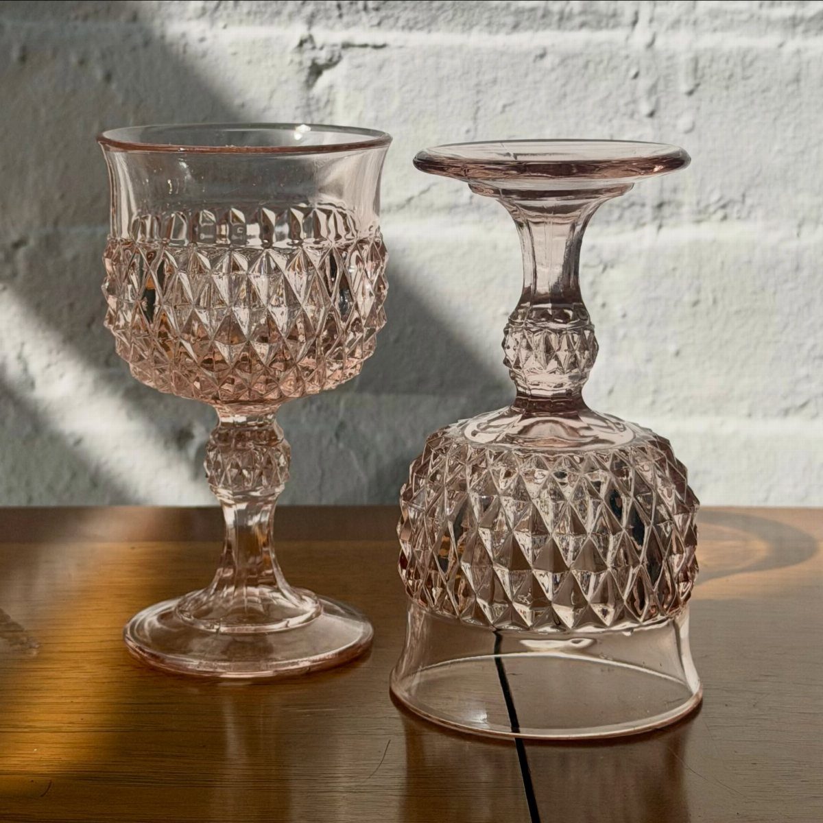 Two pink diamond-patterned glass goblets on a wooden table against a white brick wall.