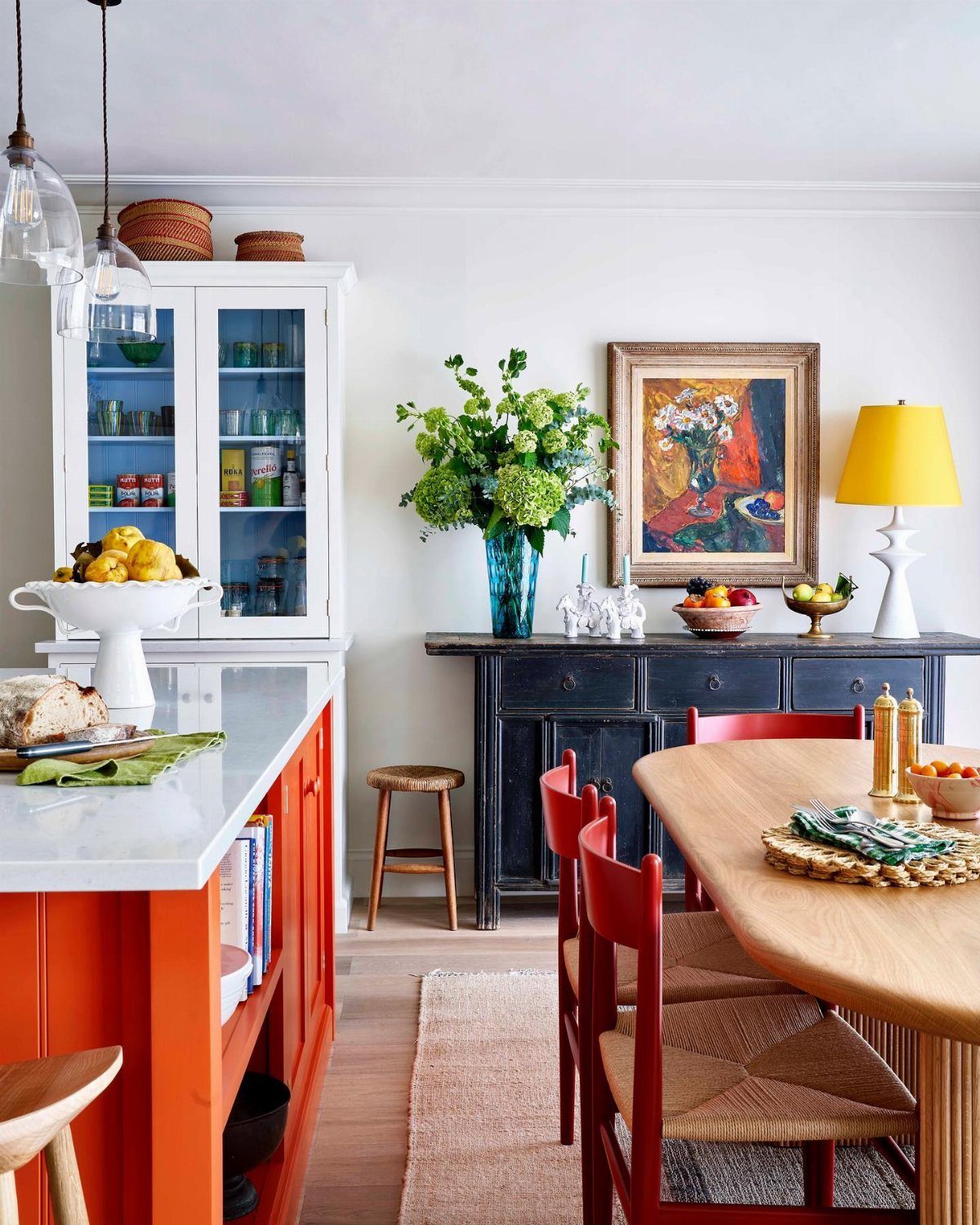 An orange kitchen island with marble top sits near a wooden dining table and red chairs.