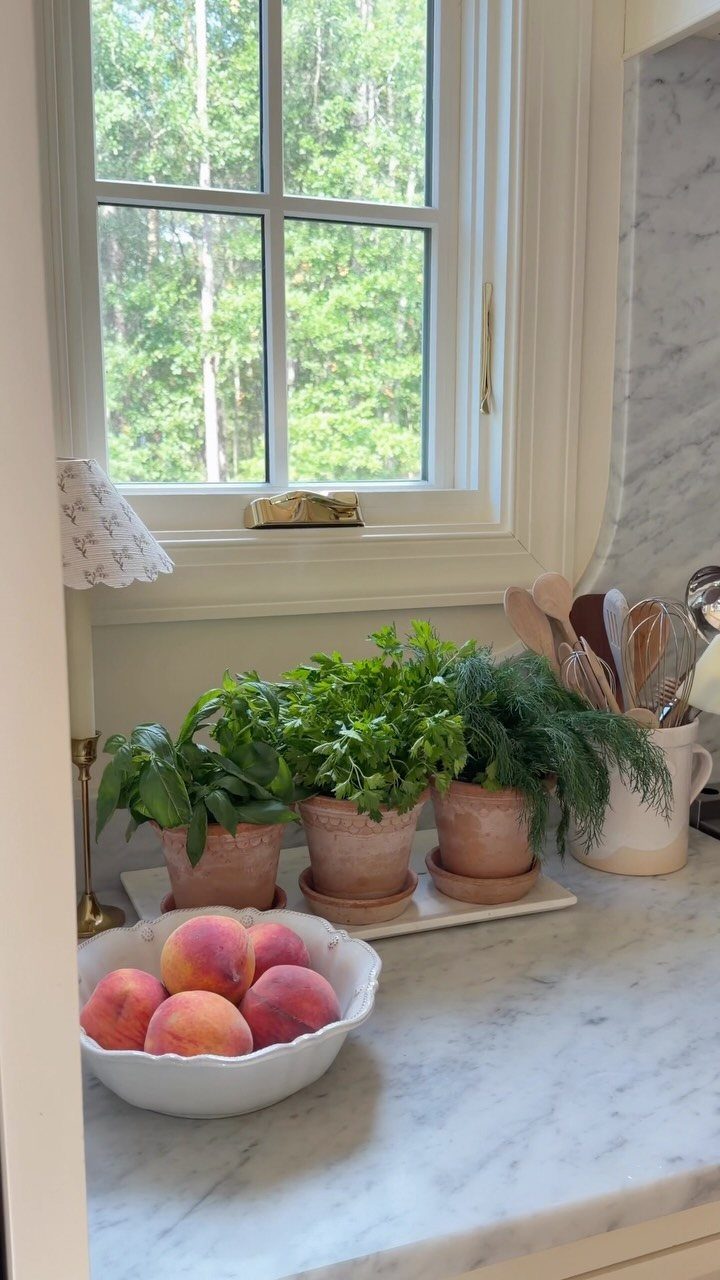 Three terracotta pots with fresh green herbs sitting on a marble kitchen countertop near a window.